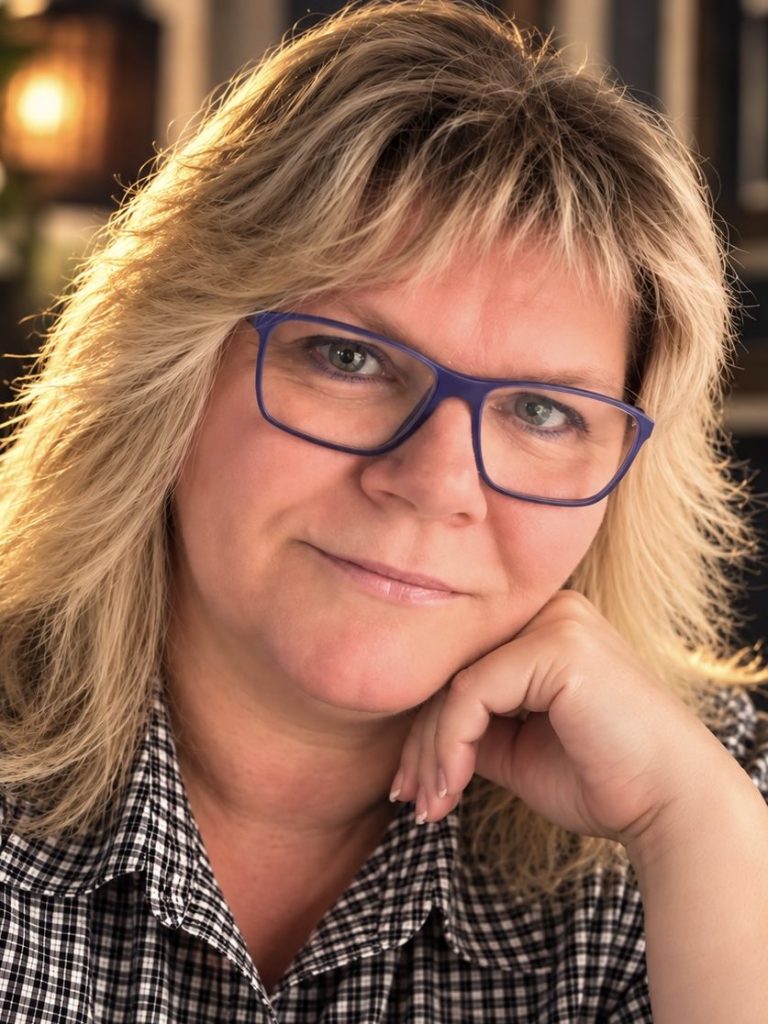 Porträt einer Frau mit blondem, leicht lockigem Haar, die lächelt. Sie trägt einen schwarzen Pullover. 